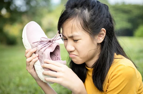 Несчастная азиатская девочка-подросток нюхает свои кроссовки, держит вонючую мотыгу в руке с отвращением, неприятный запах из-за жаркой погоды или после физических упражнений, грустная девочка с плохим запахом, накопленная грязь — стоковое фото
