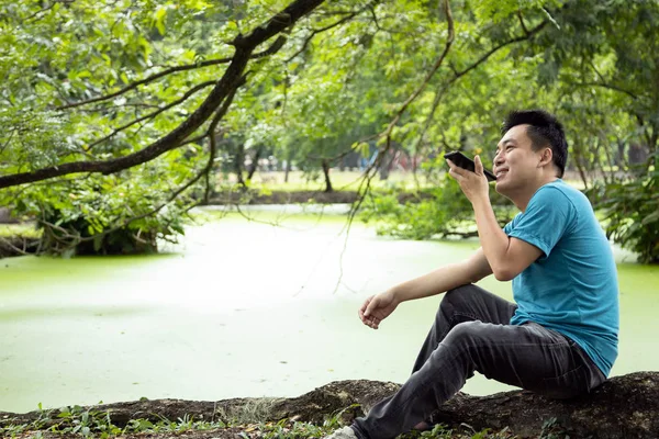Happy asian man talking on smart phone with the digital voice  assistant, sitting in green nature, male people using voice command recorder or calling on speakerphone with family, technology concept — Stock Photo, Image