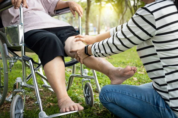Asyalı bakıcı kadın, yaşlı anne tekerlekli sandalyesinin bacağını ve dizini kontrol ediyor, yaşlı insanlar bacak ağrısı çekiyor, kızından bacak masajı alıyor, ağrıyan kaslar, fiziksel yaralanma, sağlık hizmetleri... — Stok fotoğraf