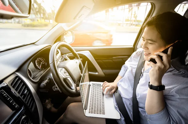 Happy asian businesswoman talking on mobile phone while  working on laptop computer in the drivers seat in her car,female people negotiating business with her clients,communication,technology concept