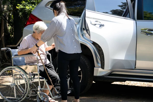 Asiática cuidador do sexo feminino ajudando a mulher idosa com deficiência em cadeira de rodas para entrar no carro, cuidado filha útil e apoiar a mãe sênior para se levantar de cadeira de rodas ao ar livre, cuidar de pessoas idosas — Fotografia de Stock