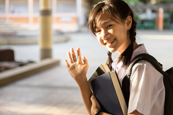 Χαμογελώντας κοριτσάκι του δημοτικού σχολείου κουβαλώντας σακίδιο πλάτης και βιβλία στο χέρι πηγαίνοντας στο σχολείο, μαθήτρια σήκωσε το δεξί της χέρι, λέγοντας γεια, χαρούμενη Ασιάτισσα έφηβη μαθήτρια με τσάντα, πίσω στο σχολείο — Φωτογραφία Αρχείου