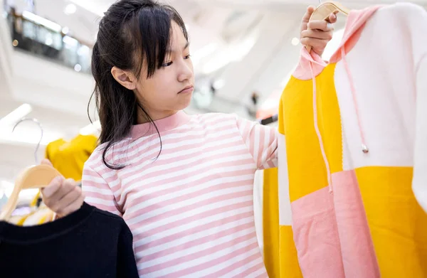 Красивая азиатская девочка ребенок выбирает одежду в магазине одежды в торговом центре, женщина подросток покупает новую моду длинные рукава футболки во время скидки для себя в качестве подарка в торговом центре — стоковое фото