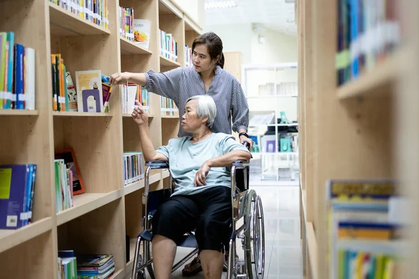 Hija asiática y madre mayor eligiendo libro en biblioteca de estantería, mujer anciana discapacitada con silla de ruedas buscando libro necesario, familia disfrutando llevando libros a la lectura, concepto de ocio — Foto de Stock