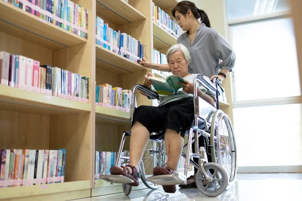 Mulher cuidadora e idosa lendo um livro na biblioteca, asiática idosos com cadeira de rodas, curtindo e lendo livros com cadeira de rodas, prevenir a depressão e perda de memória em idosos, cuidados, apoio — Fotografia de Stock