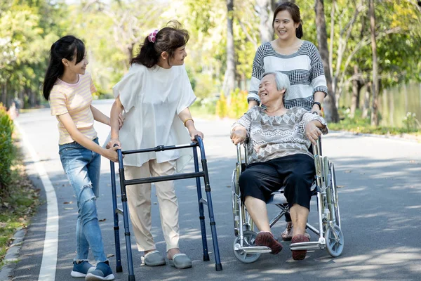 Mutlu Asyalı aile, kıdemli kadın, engelli büyükanne yürüteç ve tekerlekli sandalye, kız, genç torun açık hava parkında yürüyüş yapıyor, çocuk kız ve anne destek oluyor, yaşlılara bakıyor. — Stok fotoğraf