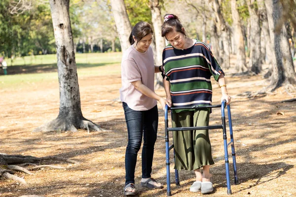 アジアのシニア女性は膝の手術後のリハビリ中に歩行補助を使用し、若いケアは安心感のある成熟した高齢者が歩行を練習するのを助け、女性介護者の助け、ケア、サポート — ストック写真