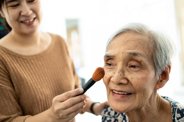 Feliz asiática mujer mayor o madre disfrutar del maquillaje en la cara, alegre mujer cuidadora o hija está ayudando a los ancianos maquillaje, aplicando rubor en la mejilla por cepillo, servicios, cuidado y apoyo para los ancianos — Foto de Stock