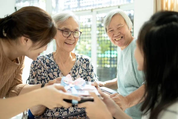 Glad asiatisk familj, dotter, barnbarn besöker och ge gåva till gamla mor- och farförälder, glada kvinnliga äldre hålla presentförpackning, kvinna, barn flicka gratulerar mormor födelsedag på vårdhem — Stockfoto