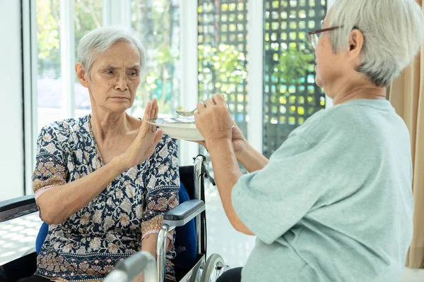 Unglückliche asiatische Seniorin ablehnend, Geste Hand nein, müde alte Menschen, die sich krank fühlen, Dysphagie, Dyspepsie oder Langeweile beim Essen, Freund füttert ältere Patientin im Rollstuhl, Appetitlosigkeit, Magersucht-Konzept — Stockfoto