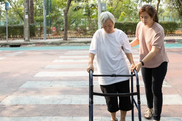 Asiatische erwachsene Tochter unterstützt und unterstützt ihre ältere Mutter beim Überqueren der Straße, ältere Frau mit Rollator auf dem Zebrastreifen, weibliche Betreuerin hilft alten Menschen im Freien beim Gehen auf dem Zebrastreifen — Stockfoto