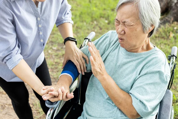 Donna anziana asiatica ferita con supporto al gomito che soffre di artrite, dolore muscolare, dolore al braccio del gomito, malattia articolare o miosite, anziana infelice con gomiti protettivi seduta sulla sedia a rotelle — Foto Stock