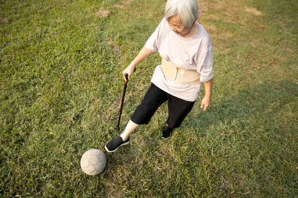 歩く棒を持つアジアの高齢者の女性は、古いサッカーで遊んで、健康的な女性の高齢者は、サッカーボールを蹴っている、身体運動、トレーニング足の筋肉、高齢者の体のバランス、医療、健康, — ストック写真