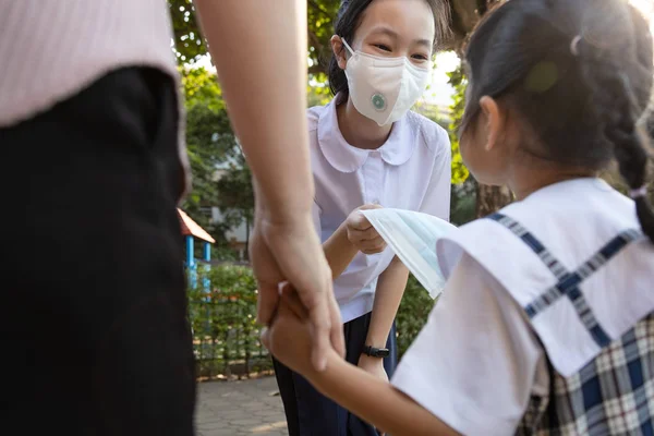 Generous girl student wearing Thai school uniform with mask,schoolgirl is sharing protection mask with asian kid girl,giving medical mask to prevent disease,coronavirus,flu season,dust,air pollution — Stok fotoğraf