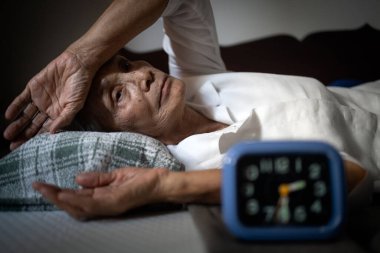 Aylardır uykusuzluk çeken yorgun, bitkin Asyalı yaşlı kadın, kronik uykusuzluk, geceleri uyuyamayan, elleri alnında ve gözleri açık, yatağında uzanmış, uykusuzluk çeken yaşlı insanlar.