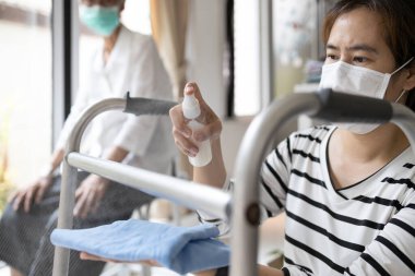 Maskeli bir bakıcı, alkol antiseptiği sıkar, dezenfekte eder, yürüyüş yardımcısını temizler, yaşlılar için yürür, Coronavirus salgını sırasında, Covid-19, evdeki yaşlı ekipmanları temizler.