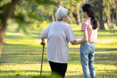 Yaşlı bir kadın yürümesine yardım etmek için baston kullanıyor. Açık havada Asyalı torunuyla dengede duruyor. Mutlu büyük anne ve gülümseyen kız el ele tutuşuyor ve birlikte yürüme egzersizleri yapıyor.