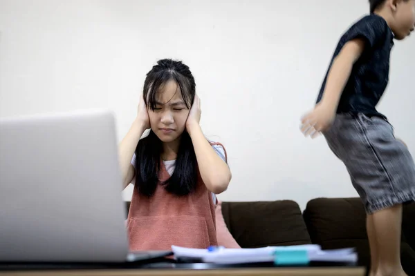 Asian child girl is using her hands to cover her ears from the loud voice,annoying noise from little brother while studying online,kid boy is jumping,play near her,problems learning online at home