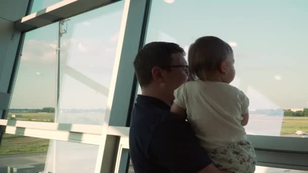 Vater und Sohn betrachten Flugzeuge durch das Glasfenster. Reise mit Baby am Flughafen — Stockvideo