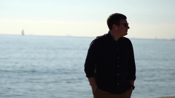 Exitoso hombre caucásico escuchando música en auriculares inalámbricos en la playa del mar. Nómada digital — Vídeo de stock