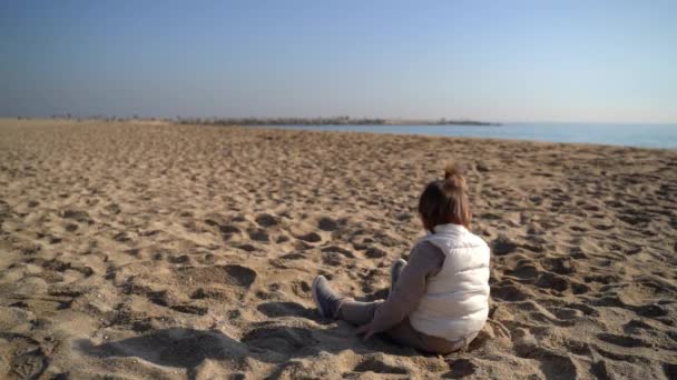 Soğuk rüzgarlı bir günde kumsalda bebek. Kıyı şeridinde oturuyor. Kumla oynuyorum. İspanya — Stok video