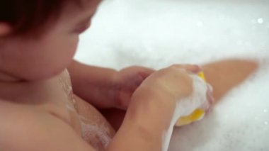 Küçük kız banyoda köpükle ve sarı plastik ördekle yıkanıyor.
