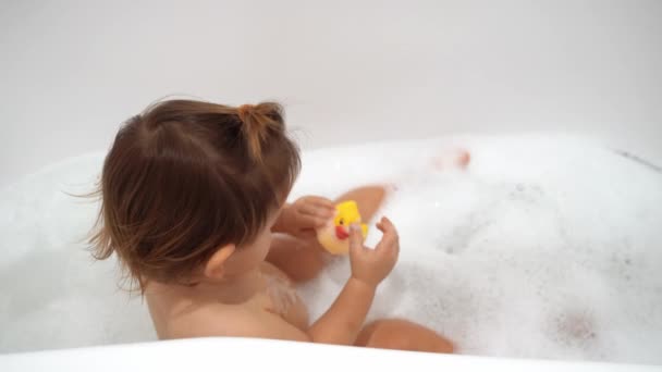 Bebé niña lavándose en el baño con espuma y pato de goma amarillo — Vídeo de stock