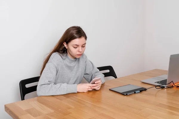 En tonårstjej sitter vid träbordet och förhalar i sociala medier. Problem med missbruk — Stockfoto