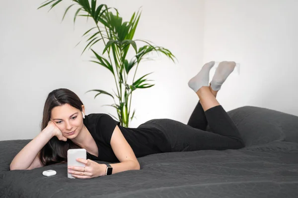 Egy fiatal nő telefonnal és vezeték nélküli fülhallgatóval az ágyon. Maradj otthon a karantén alatt. — Stock Fotó
