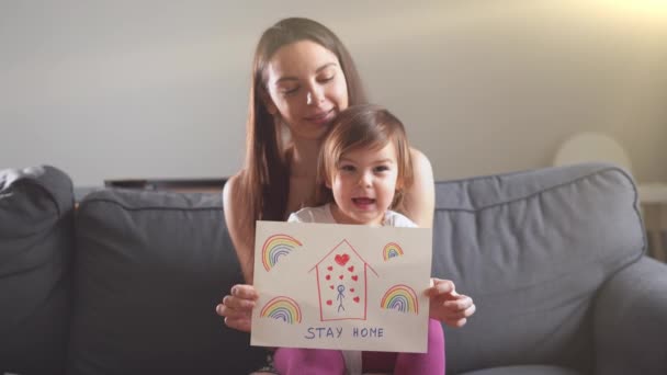 Glückliche junge Mutter und Baby auf dem Sofa mit Zeichnung STAY HOME und Regenbogen. Globale Kampagne für COVID 19 — Stockvideo