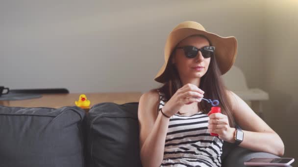 Молода жінка в приміщенні вдома дме мильні бульбашки, прикидаючись на пляжі — стокове відео