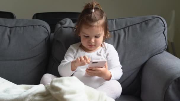 Κοριτσάκι που κάθεται στον καναπέ στο σπίτι και παίζει παιχνίδια στο smartphone — Αρχείο Βίντεο