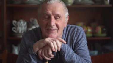80 'li yaşlarda yaşlı bir adam oturmuş elinde sopayla kamerayla konuşuyor.