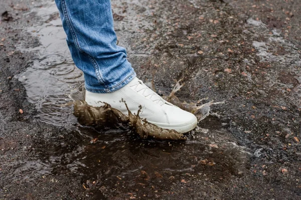 Botas blancas en un charco fangoso en una carretera asfaltada. Primer plano. Rocía desde las botas . Fotos De Stock