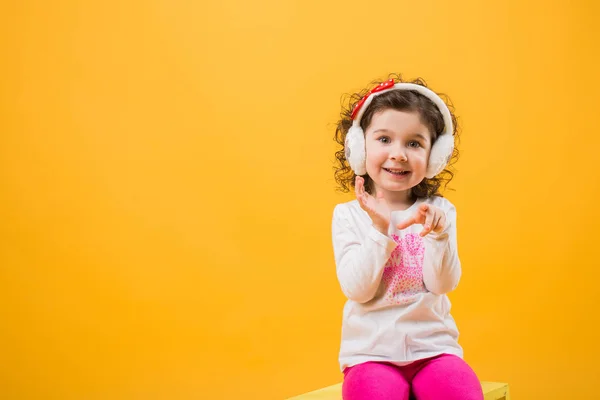 Pequeña alegre chica hermosa feliz de pie aislado sobre un fondo amarillo en auriculares suaves, emociones infantiles . —  Fotos de Stock