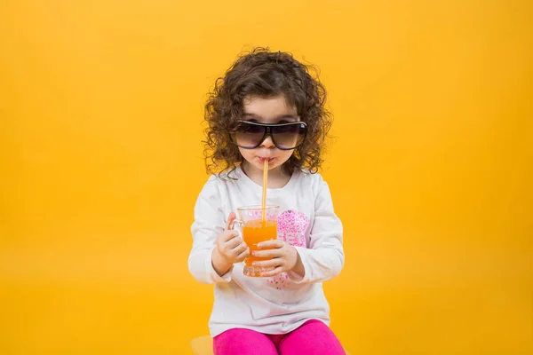 Niña en un vaso bebe jugo de naranja sobre un fondo amarillo. Emoción infantil, humor, espacio para el texto . —  Fotos de Stock