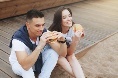 Genç ve pozitif bir çift sahilde oturmuş hamberger yiyor. Yaşam tarzı, aşk ve fast food.