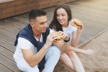 Genç ve pozitif bir çift sahilde oturmuş hamberger yiyor. Yaşam tarzı, aşk ve fast food.