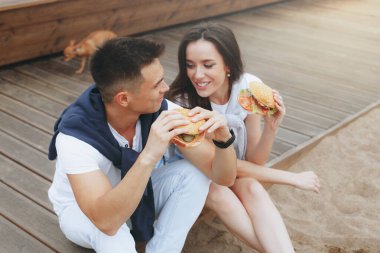 Genç ve pozitif bir çift sahilde oturmuş hamberger yiyor. Yaşam tarzı, aşk ve fast food.