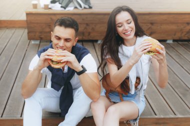 Genç ve pozitif bir çift sahilde oturmuş hamberger yiyor. Yaşam tarzı, aşk ve fast food.
