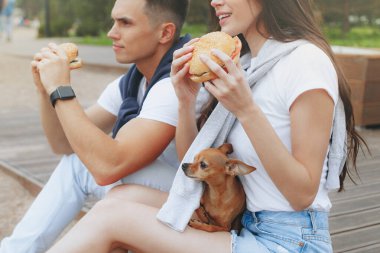 Genç ve pozitif bir çift sahilde oturmuş hamberger yiyor. Yaşam tarzı, aşk ve fast food.