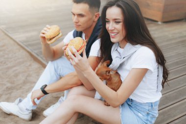 Genç ve pozitif bir çift sahilde oturmuş hamberger yiyor. Yaşam tarzı, aşk ve fast food.