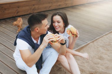 Genç ve pozitif bir çift sahilde oturmuş hamberger yiyor. Yaşam tarzı, aşk ve fast food.