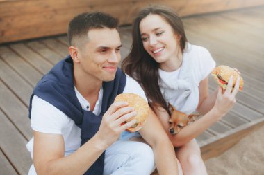 Genç ve pozitif bir çift sahilde oturmuş hamberger yiyor. Yaşam tarzı, aşk ve fast food.