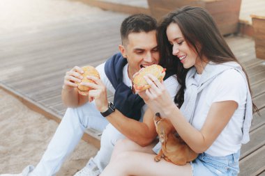 Genç ve pozitif bir çift sahilde oturmuş hamberger yiyor. Yaşam tarzı, aşk ve fast food.