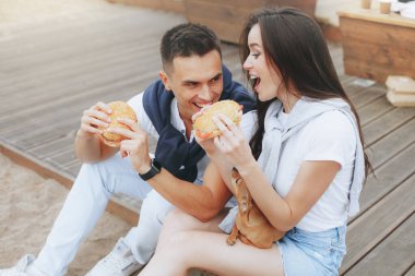 Genç ve pozitif bir çift sahilde oturmuş hamberger yiyor. Yaşam tarzı, aşk ve fast food.
