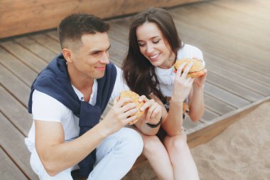 Genç ve pozitif bir çift sahilde oturmuş hamberger yiyor. Yaşam tarzı, aşk ve fast food.