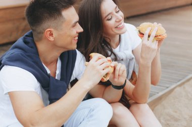 Genç ve pozitif bir çift sahilde oturmuş hamberger yiyor. Yaşam tarzı, aşk ve fast food.