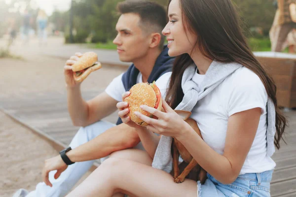 Genç ve pozitif bir çift sahilde oturmuş hamberger yiyor. Yaşam tarzı, aşk ve fast food.