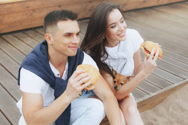 Genç ve pozitif bir çift sahilde oturmuş hamberger yiyor. Yaşam tarzı, aşk ve fast food.
